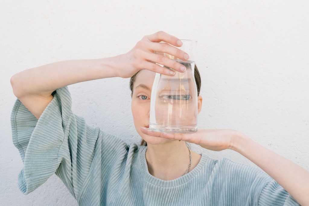 A woman holding a glass of water creating a unique facial reflection in a minimalist setting.